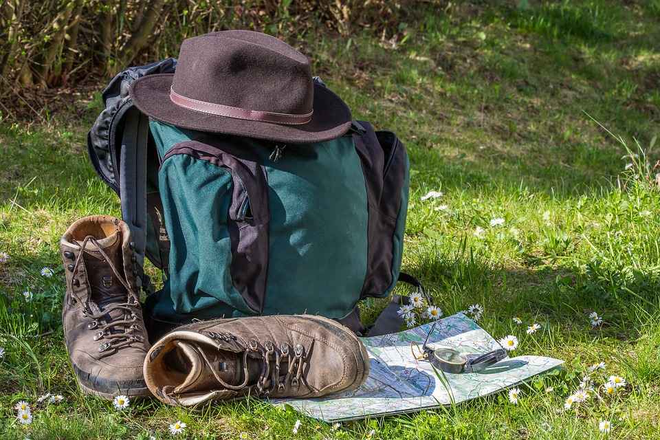 Zelený batoh na výšlap s kloboukem , botami, mapou a buzolou v trávě